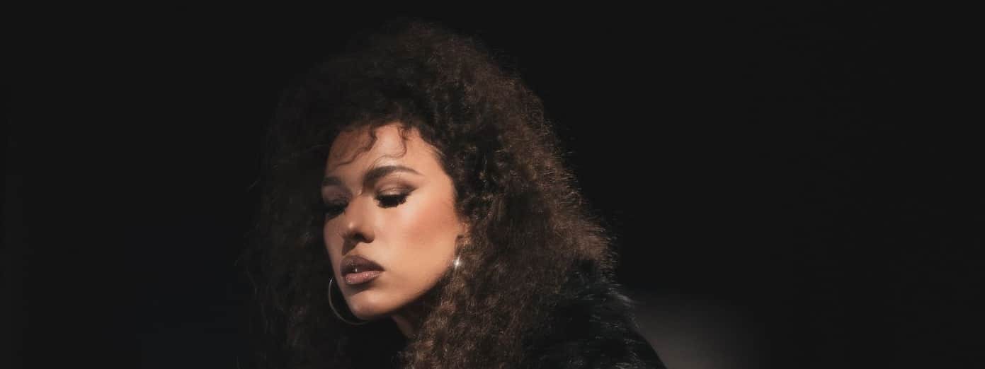 Person with curly dark hair in black leather jacket against dark background, gazing downward.