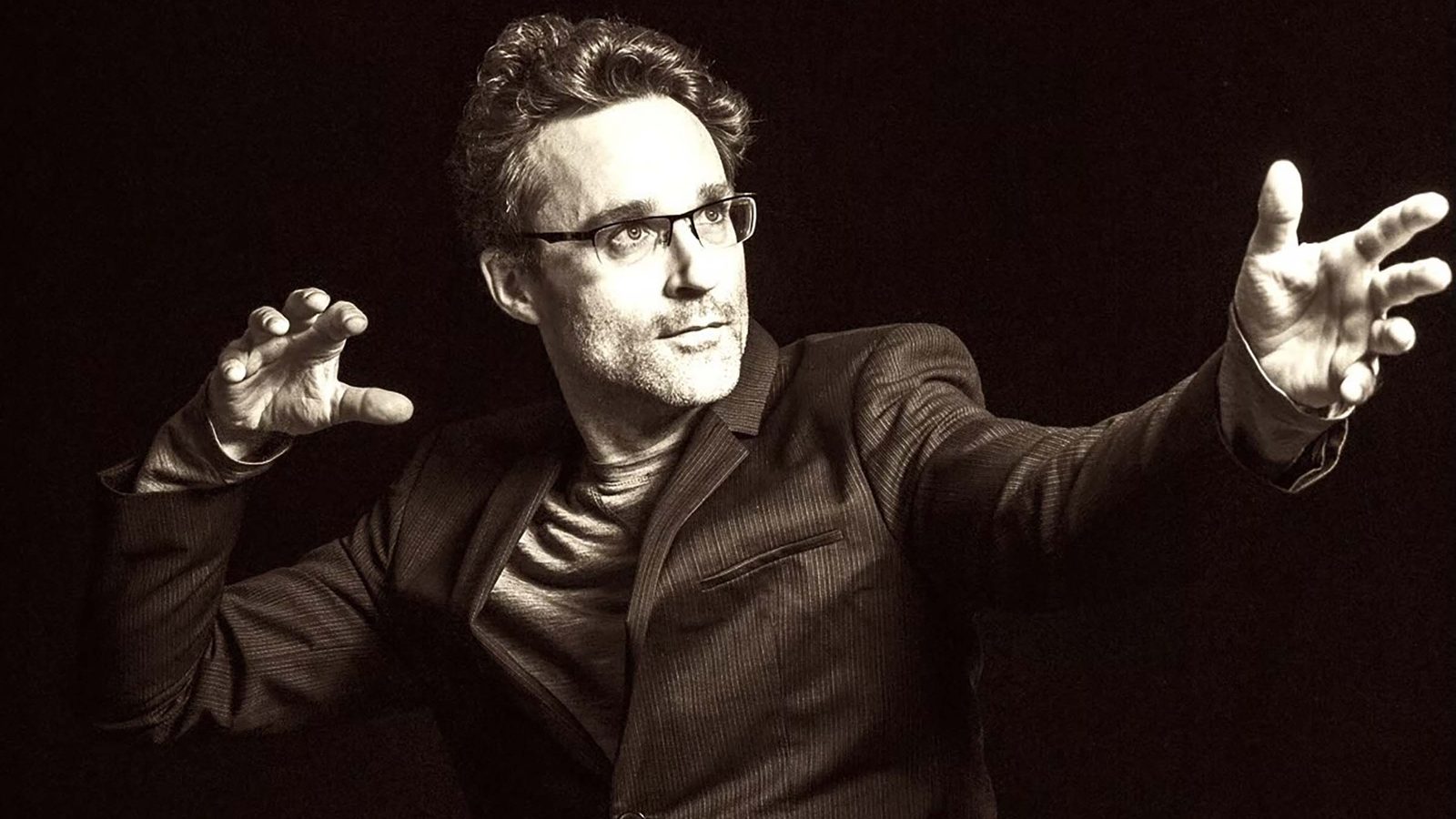 Man in dark suit and glasses gestures expressively against black background during performance.