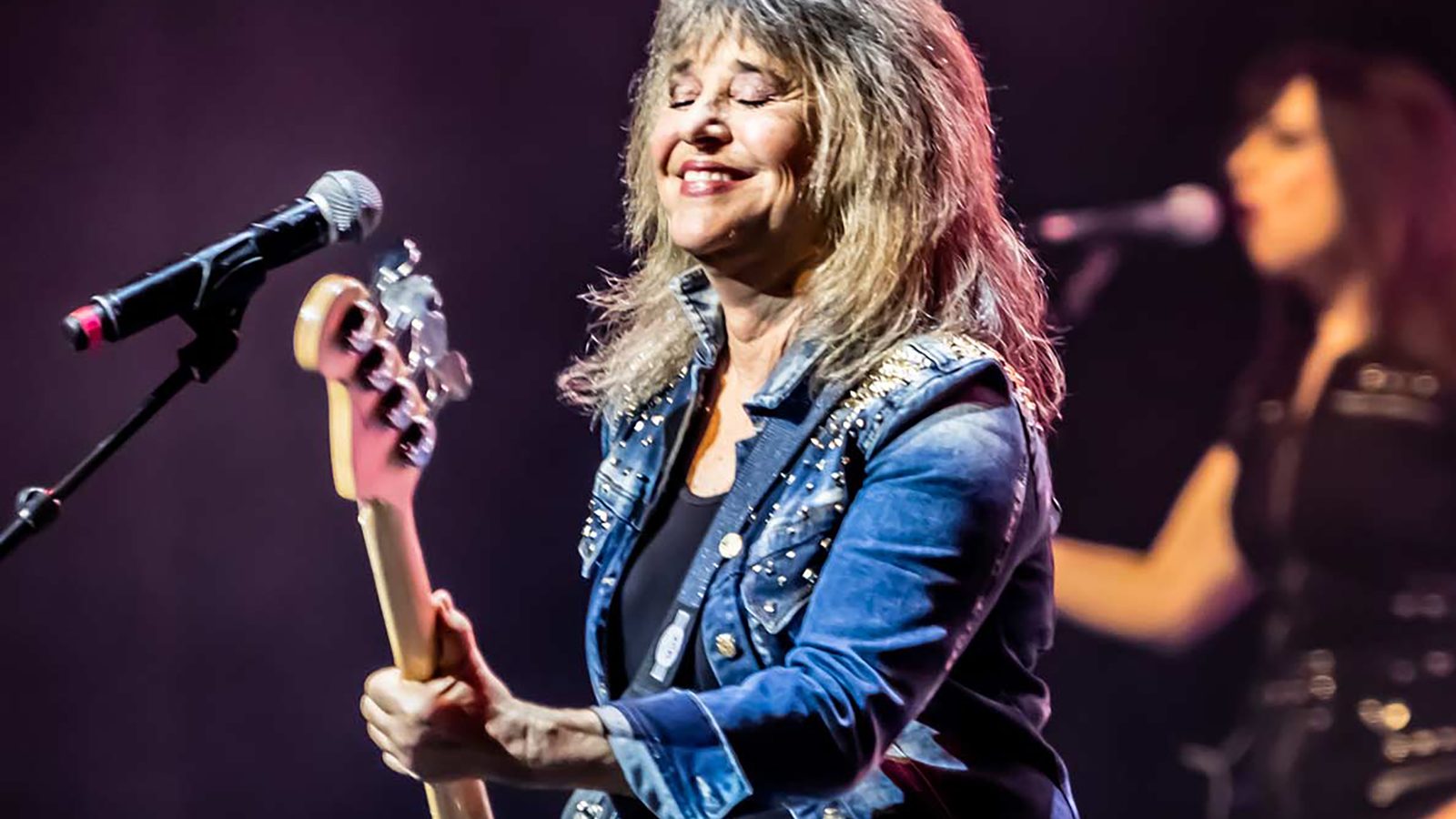 SUZI QUATRO - Musician in a blue jacket plays bass guitar at a microphone on stage with a band member visible in the background.