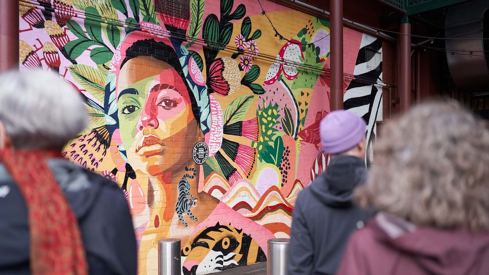 ,000 artists take over South Australia in August's SALA Festival - Gallery visitors view a vibrant portrait mural featuring a face painted in pink, green and yellow tones surrounded by botanical imagery.
