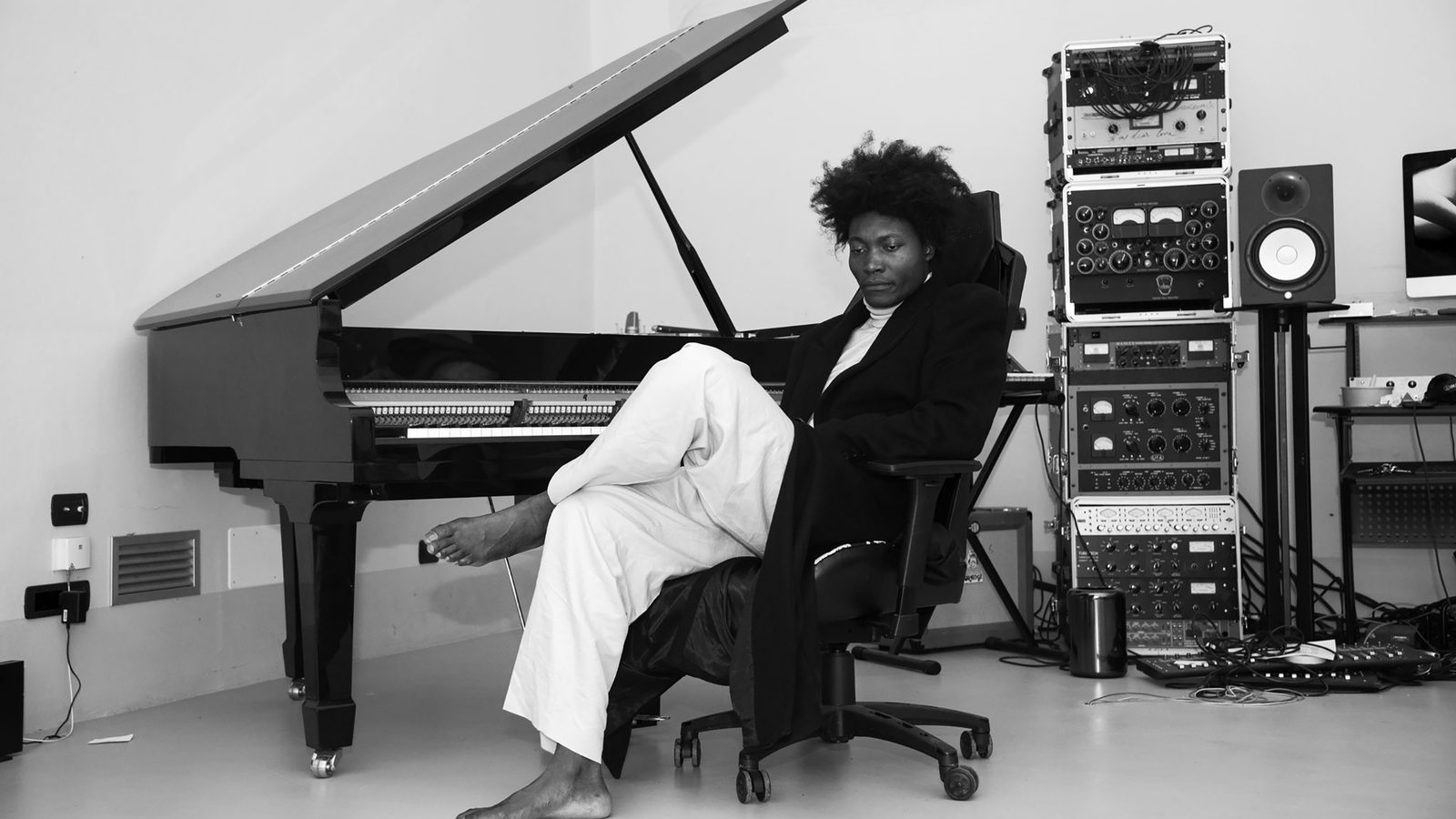 Benjamin Clementine - Musician seated at grand piano in recording studio with stacked amplifiers and equipment visible against white wall.