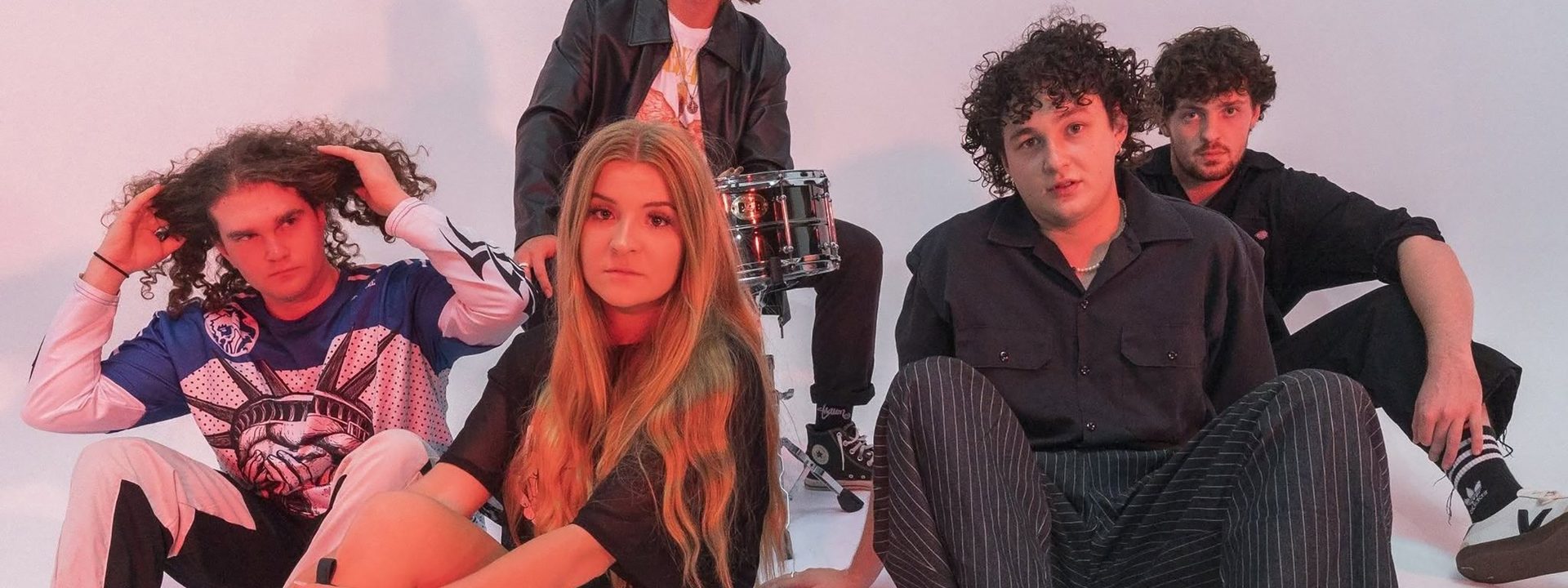 Five band members posed together against a white background lit by pink and red stage lighting.