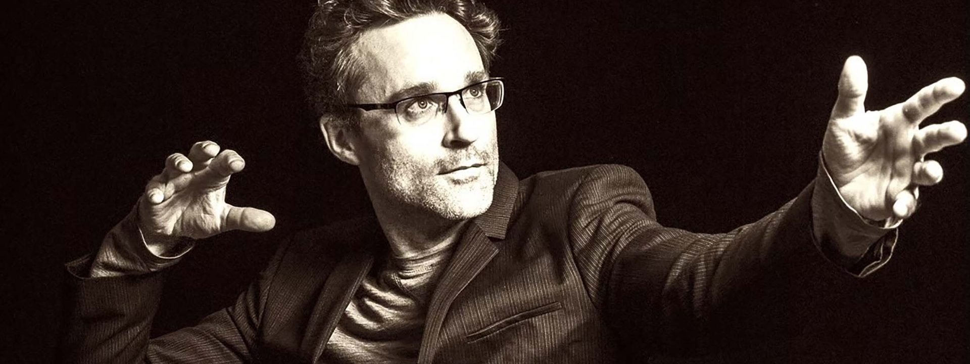 Man in dark suit and glasses gestures expressively against black background during performance.