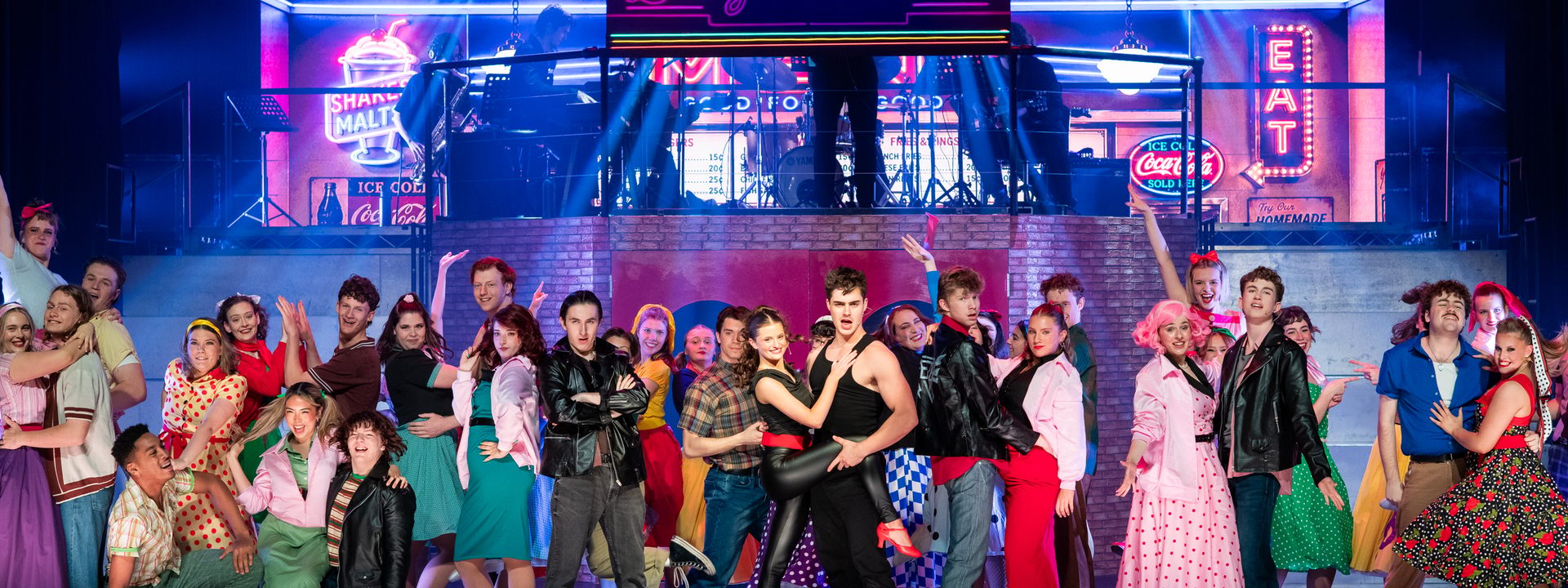 Large ensemble cast in colourful 1950s costumes performing on a brightly lit stage during Grease production.