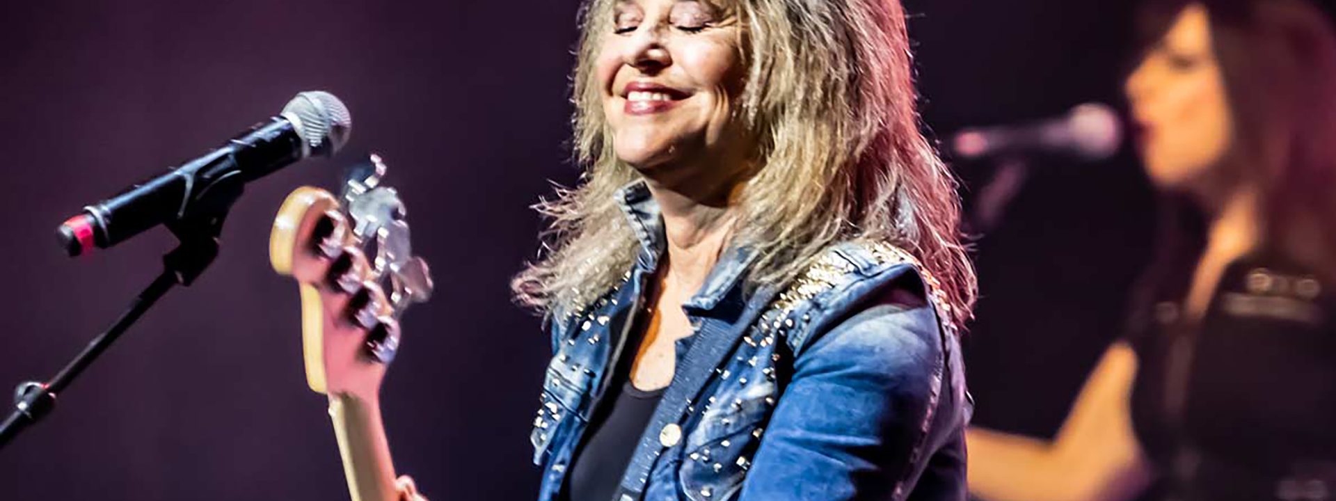 SUZI QUATRO - Musician in a blue jacket plays bass guitar at a microphone on stage with a band member visible in the background.
