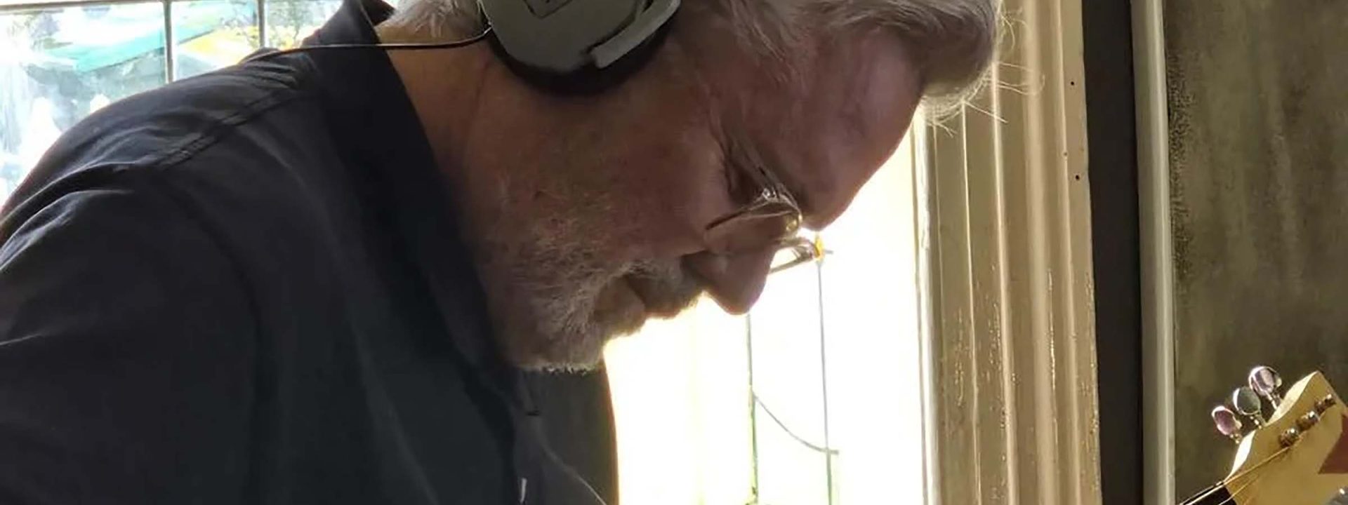 Musician with headphones around neck playing acoustic guitar indoors near a window.
