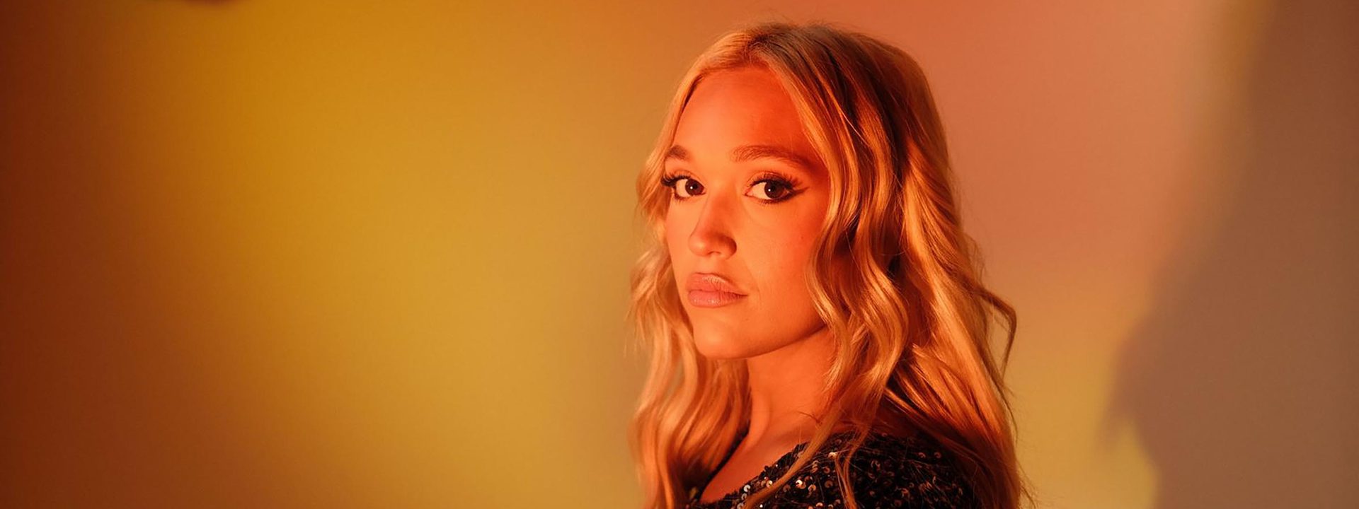 IVANA remakes Jennifer Paige and Cascada for the dancefloor - Performer with long blonde hair in a sequined black jacket against warm orange and yellow stage lighting.