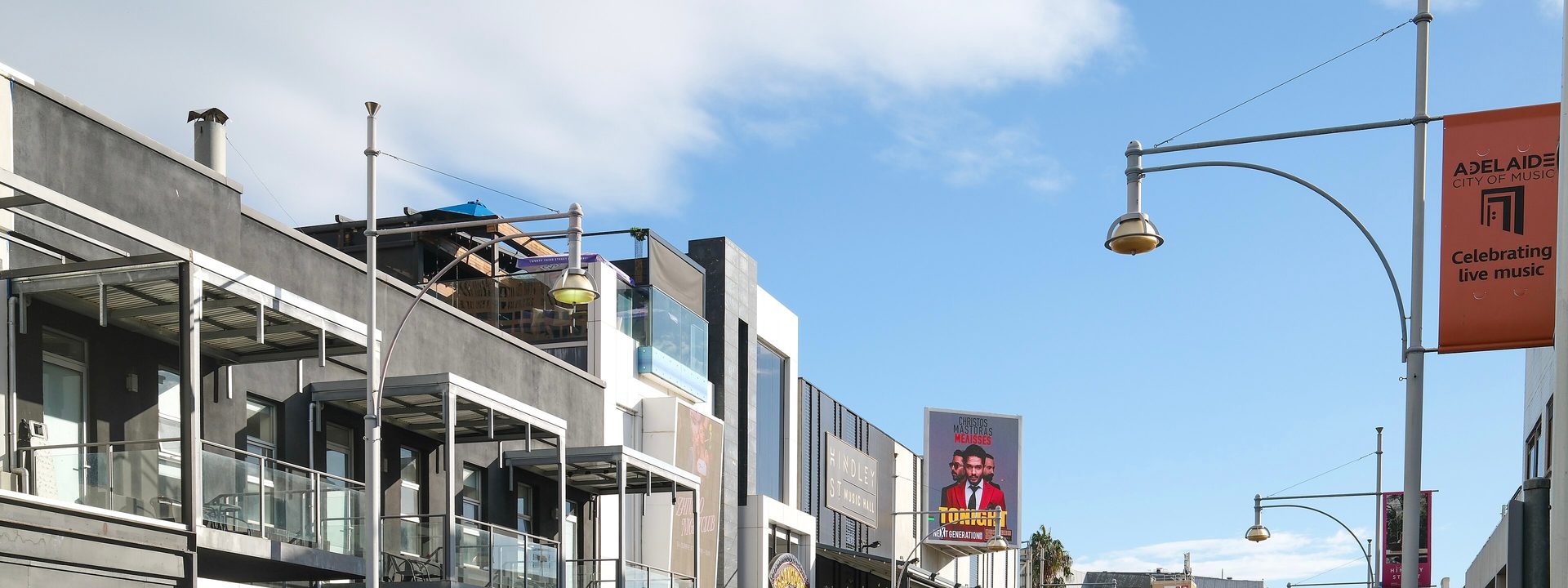 Hindley Street Music Hall - A street view of a commercial district with multi-storey buildings, shops, and parked cars under blue sky.
