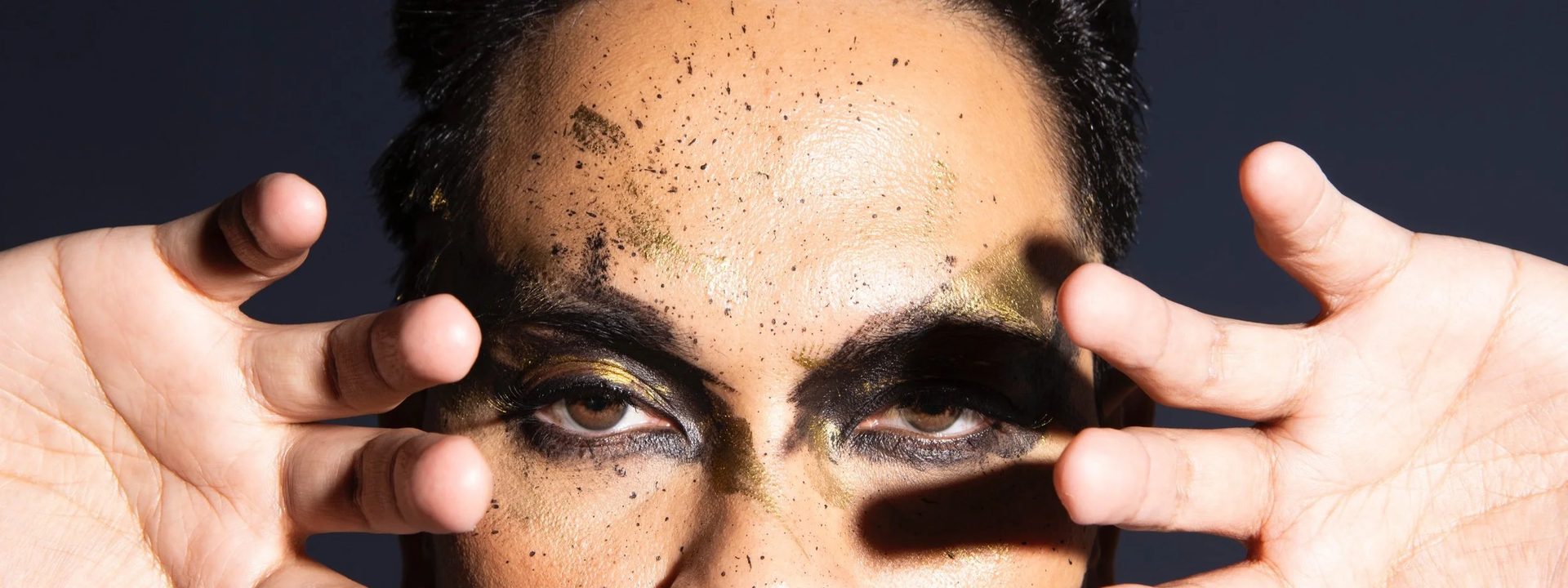 Carl De Villa takes on Prince's purple legacy at Lion Arts Factory - Performer with hands framing face, wearing ornate jewelled jackets and bold eye makeup against a dark blue background.