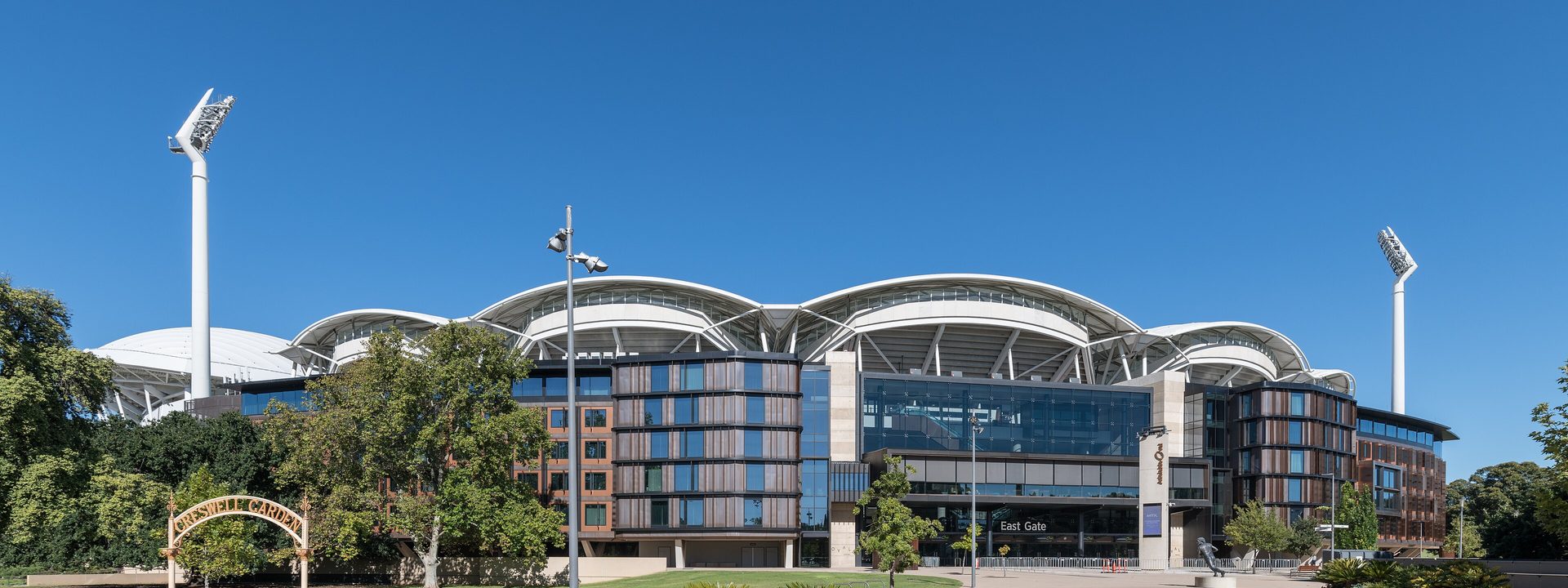 Adelaide Oval - Modern curved building with blue glass facade, landscaped courtyard, and tall white flagpoles under clear sky.