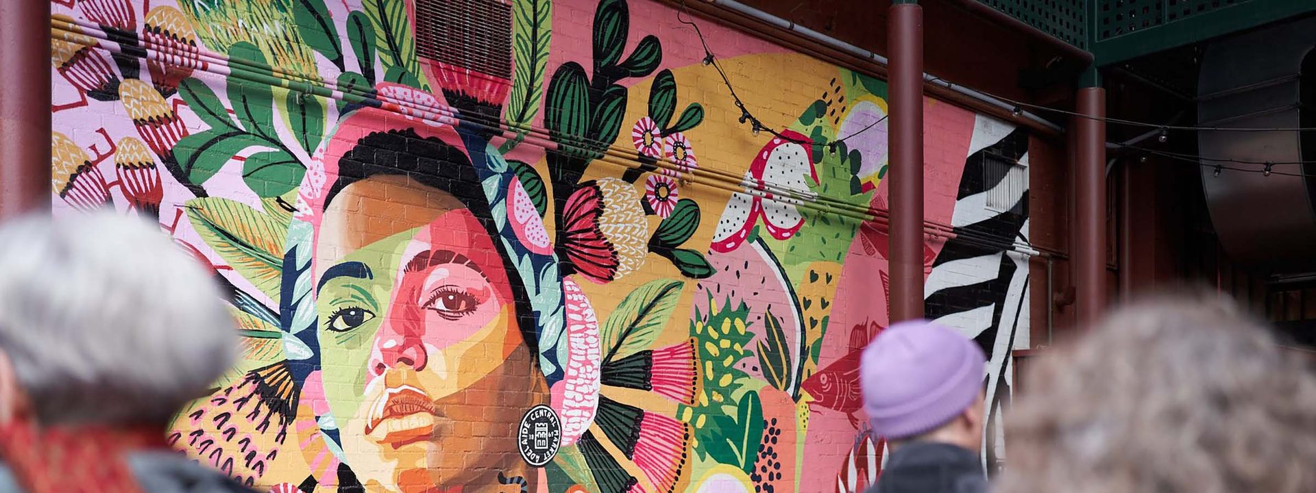 ,000 artists take over South Australia in August's SALA Festival - Gallery visitors view a vibrant portrait mural featuring a face painted in pink, green and yellow tones surrounded by botanical imagery.
