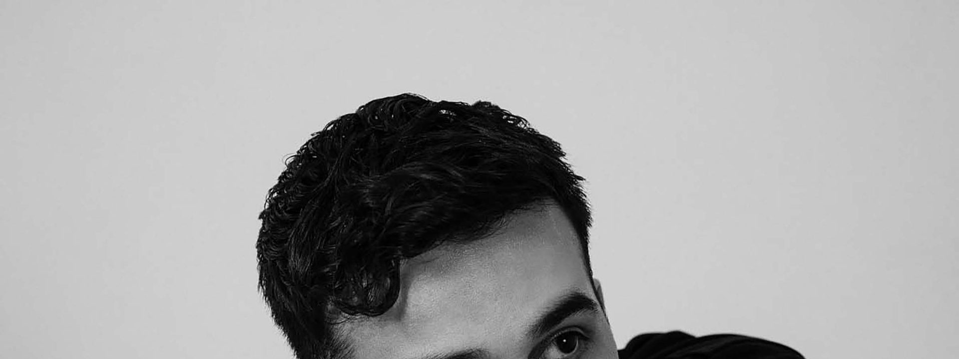 Man with dark hair resting his chin on his arms against a neutral grey background in black and white portrait.