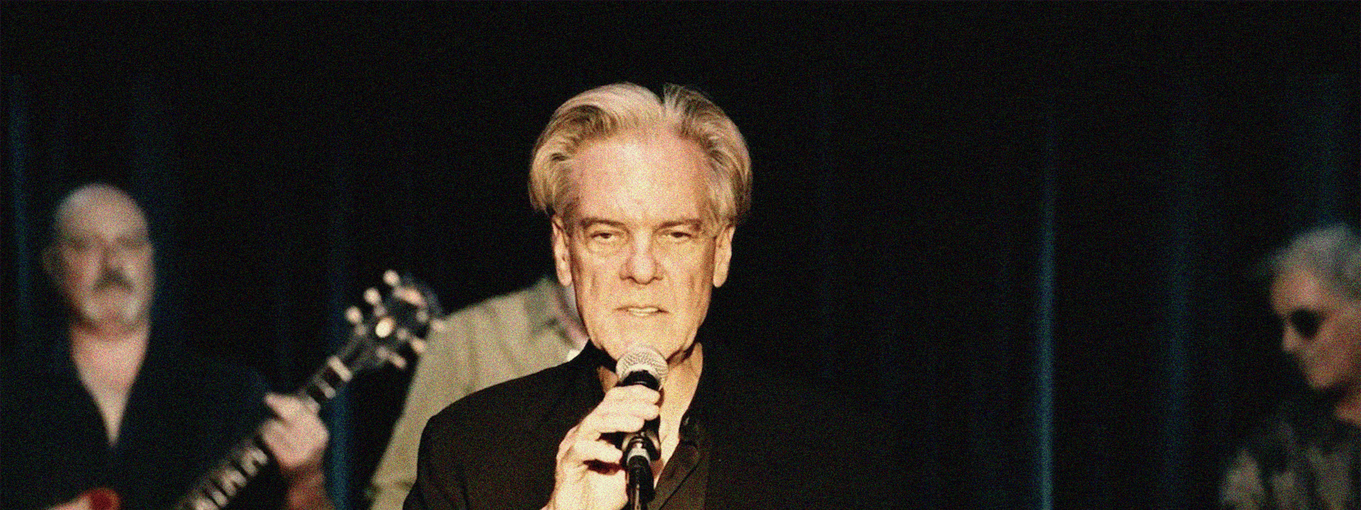 Older male singer in dark suit holding microphone centre stage, with band members visible either side.
