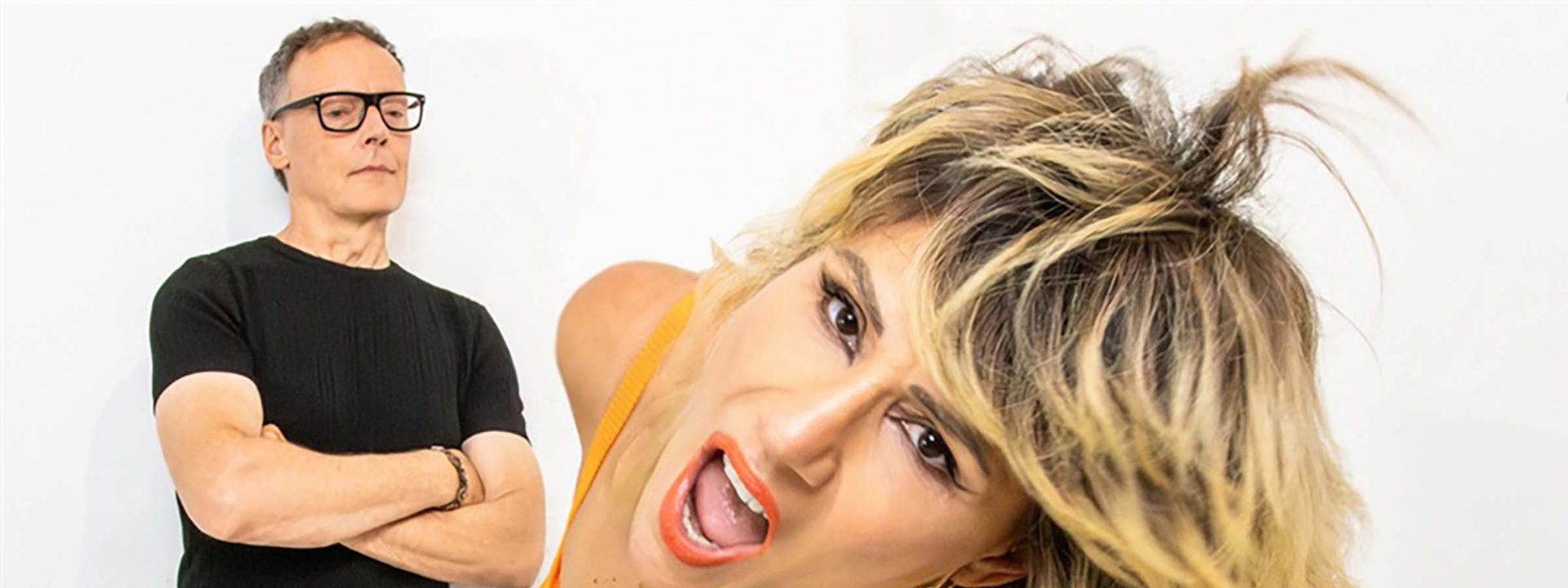 Two people posed against a white background: a man in black shirt and glasses with arms crossed, and a woman in an orange top with blonde hair and gold jewellery.
