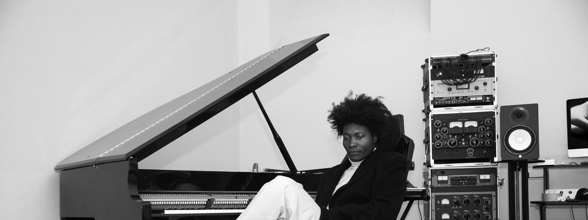 Benjamin Clementine - Musician seated at grand piano in recording studio with stacked amplifiers and equipment visible against white wall.