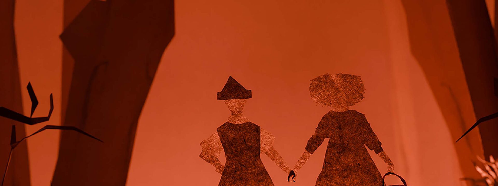 Two silhouetted figures in period costume stand together against a warm orange backdrop in a stylised theatrical scene.