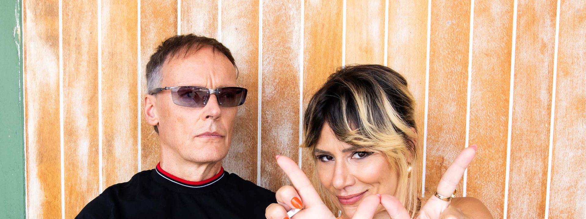Two people pose playfully against a wooden backdrop, making hand gestures toward the camera.