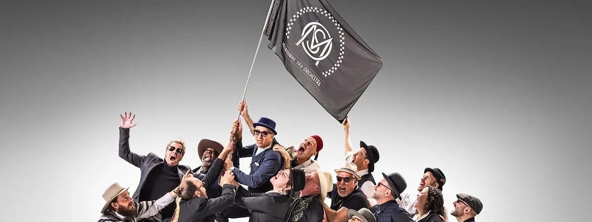 Group of performers in dark suits clustered together, one holding aloft a grey flag, against a neutral background.