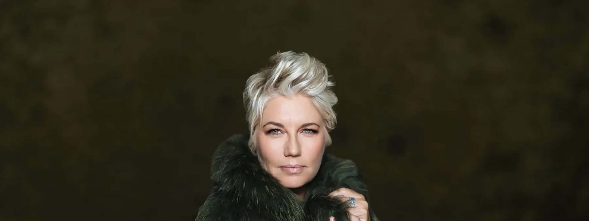 Person with platinum blonde hair wearing a black fur-collared coat against a dark background.