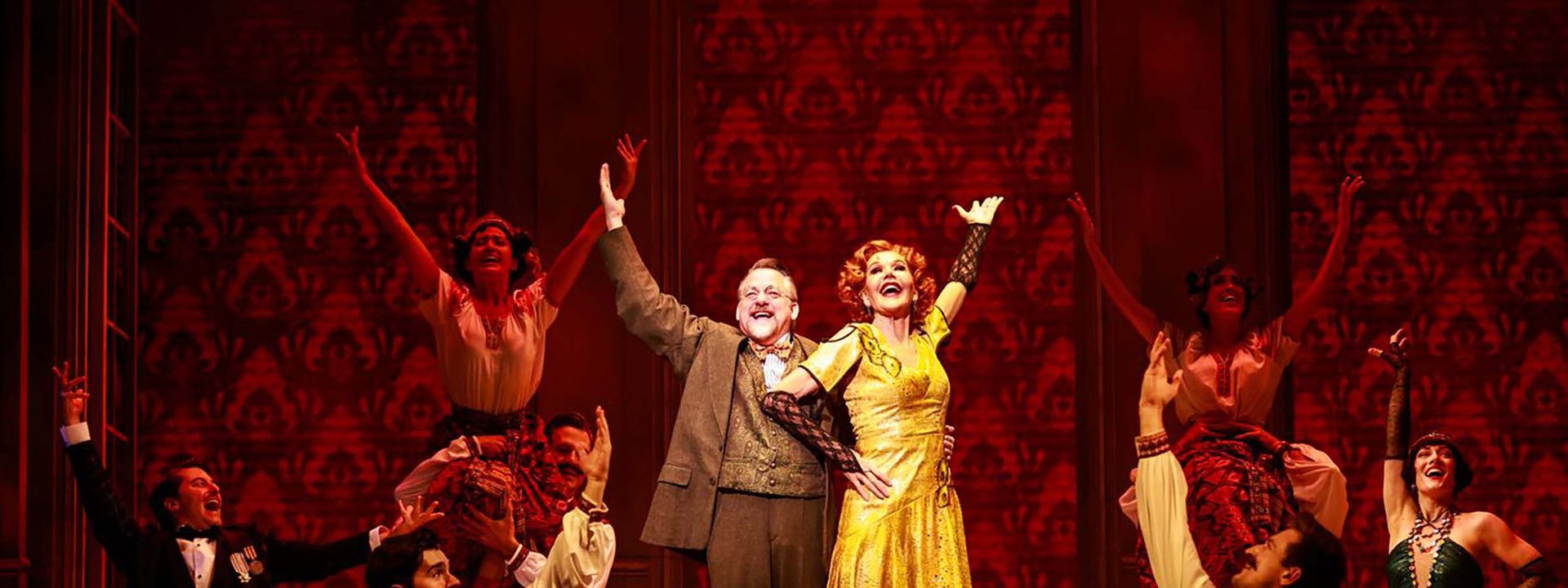 Two performers in vintage clothing stand centre stage with arms raised, surrounded by ensemble cast on a richly patterned red set.