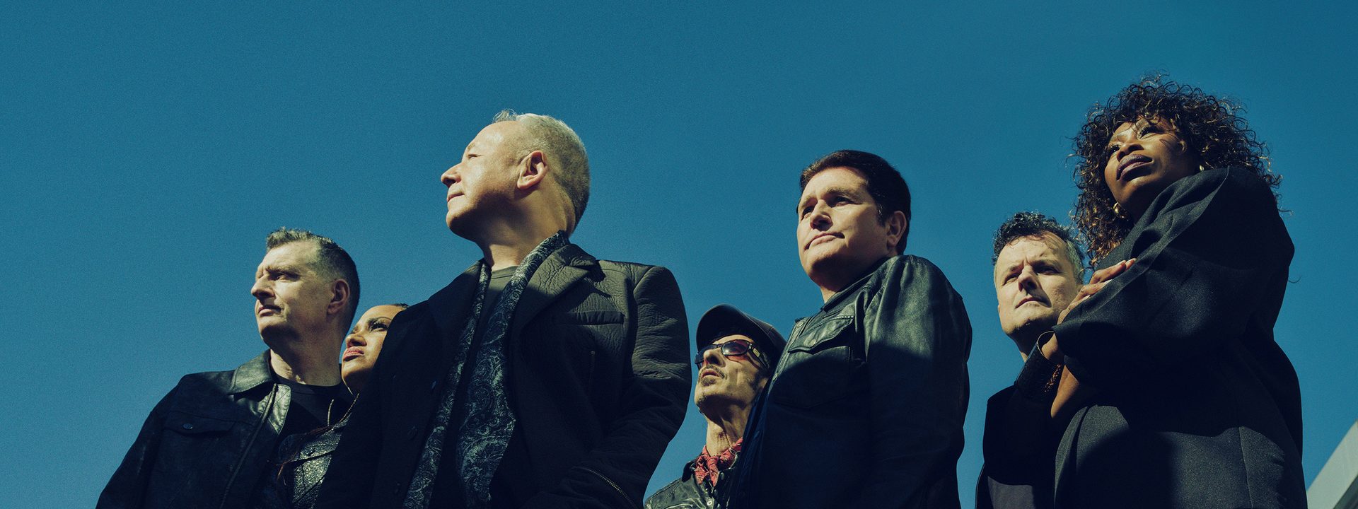 Five band members in dark suits stand against a blue sky, looking upward in a promotional pose.
