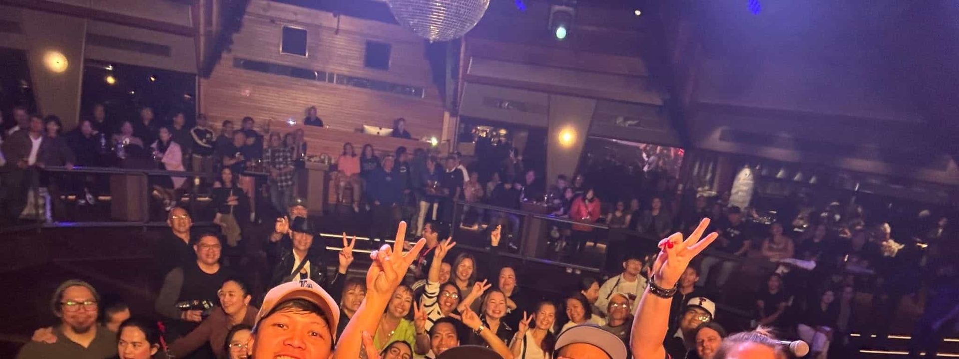 Crowd of enthusiasts in 90s attire dancing and cheering with arms raised at a concert venue under red stage lighting.