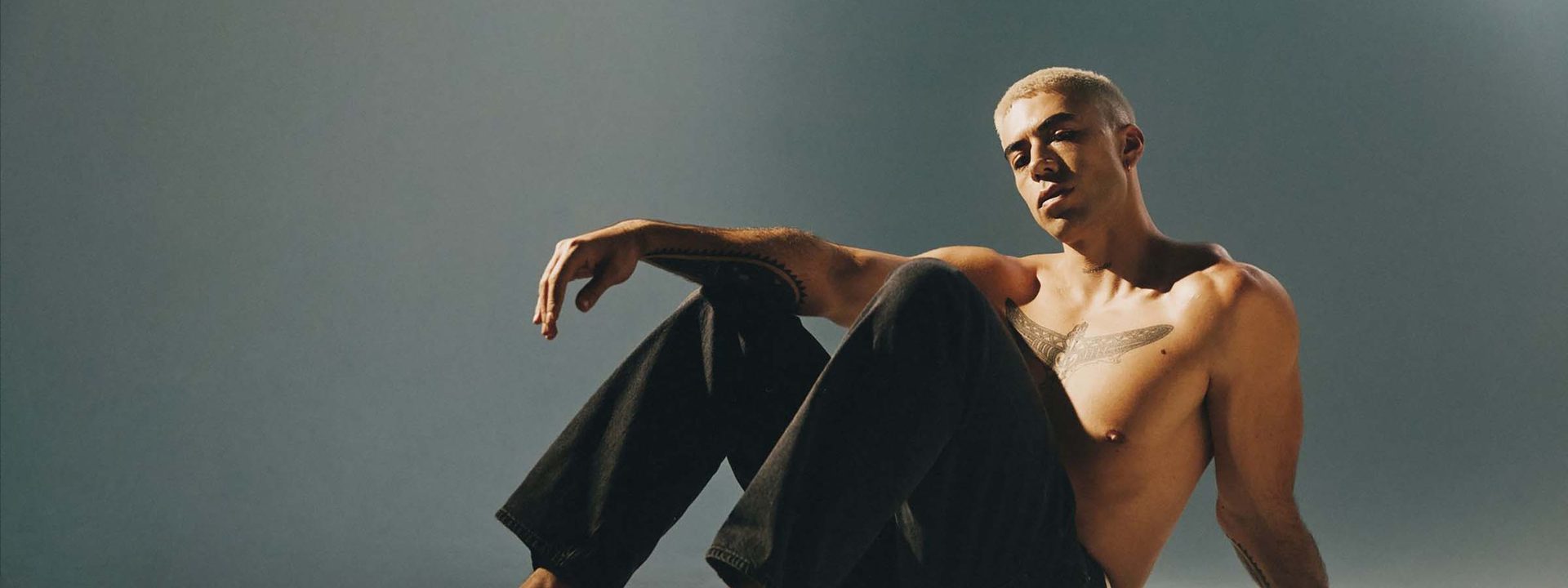 Teeks - Shirtless man seated on floor against grey background, wearing dark trousers and glasses.