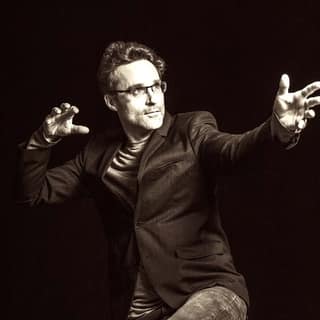Man in dark suit and glasses gestures expressively against black background during performance.