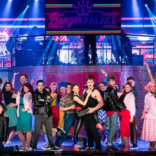 Large ensemble cast in colourful 1950s costumes performing on a brightly lit stage during Grease production.