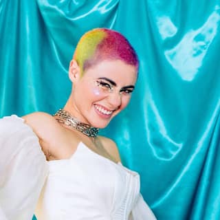 Montaigne - Glorious Heights 10th Anniversary Tour - Person with vibrant rainbow hair and gold chain jewellery against a turquoise fabric backdrop.
