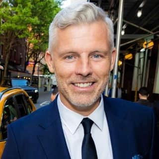 Irish comic Des Bishop finally tours Australia, stopping at Norwood in November - Man in navy suit and tie smiling at camera on a city street with yellow taxi and buildings behind.