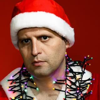 Adam Kay brings hospital Christmas chaos to Norwood Concert Hall - Man in Santa hat and tinsel garland against red background, serious expression.