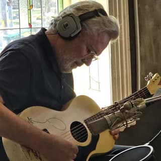 Musician with headphones around neck playing acoustic guitar indoors near a window.