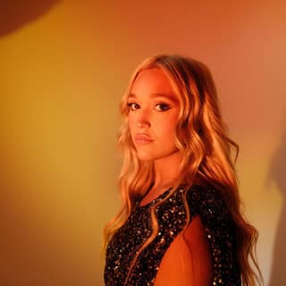 IVANA remakes Jennifer Paige and Cascada for the dancefloor - Performer with long blonde hair in a sequined black jacket against warm orange and yellow stage lighting.