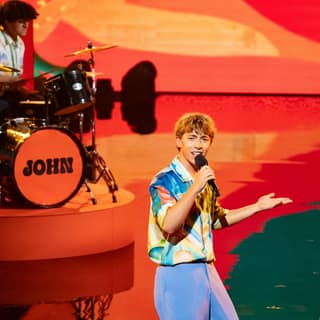 Johnny Jukebox - John Standley Live at Norwood Hotel! - Singer in blue and white shirt performing with microphone as drummer plays on colourful stage backdrop.