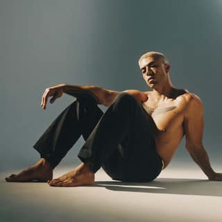 Teeks - Shirtless man seated on floor against grey background, wearing dark trousers and glasses.