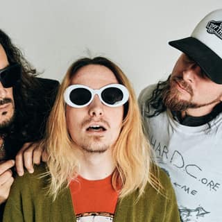 NIRVANNA [USA} - Tribute To Nirvana - Three musicians in 1990s-style clothing and sunglasses pose against a white background for NIRVANNA tribute band.