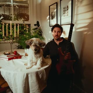 Plini ‘An Unnameable Desire’ Australia & New Zealand Tour - Musician sits at a table with a small dog in a warmly lit interior space with plants and framed artwork.