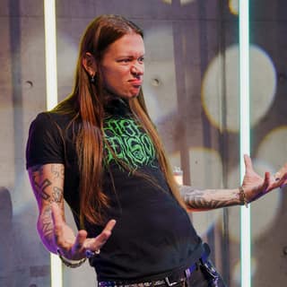 Nat’s What I Reckon - Performer with long brown hair and tattooed arms gestures expressively on stage during Nat's What I Reckon show.
