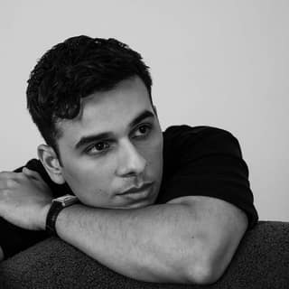 Man with dark hair resting his chin on his arms against a neutral grey background in black and white portrait.