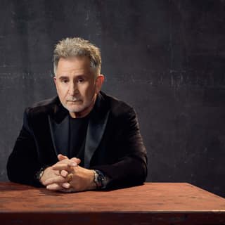 Person with blonde hair in black blazer seated at wooden table against dark textured background.