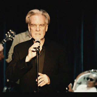 Older male singer in dark suit holding microphone centre stage, with band members visible either side.