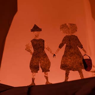 Two silhouetted figures in period costume stand together against a warm orange backdrop in a stylised theatrical scene.