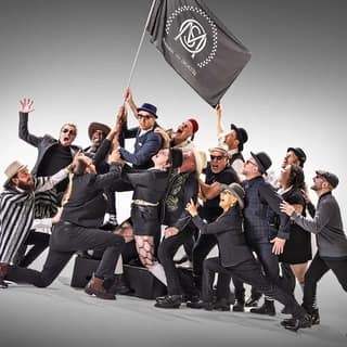 Group of performers in dark suits clustered together, one holding aloft a grey flag, against a neutral background.