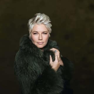 Person with platinum blonde hair wearing a black fur-collared coat against a dark background.