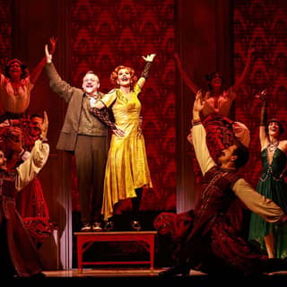 Two performers in vintage clothing stand centre stage with arms raised, surrounded by ensemble cast on a richly patterned red set.