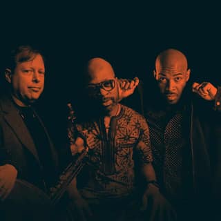 Five male musicians pose together holding instruments against a dark background.