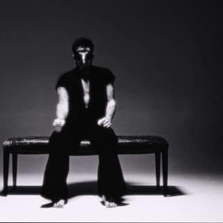 Person in black and white outfit sitting on a bench against a stark grey backdrop.