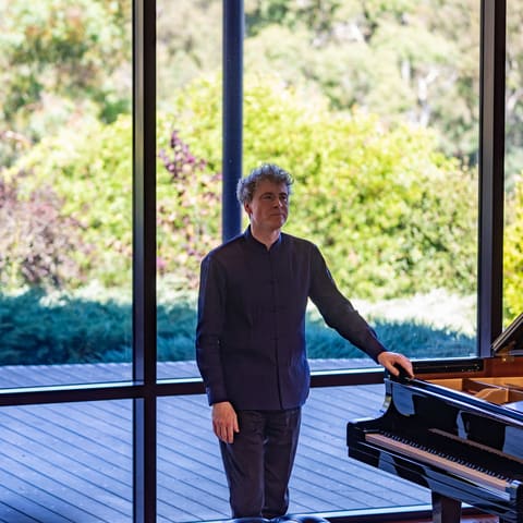 Paul Lewis - Pianist in black attire standing beside a grand piano on a wooden stage with expansive windows overlooking leafy gardens.