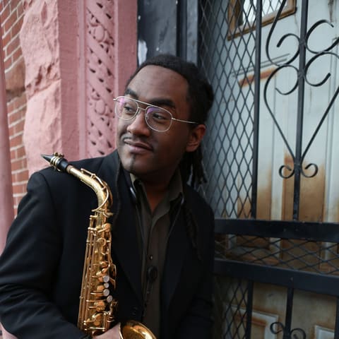 James Morrison Concert Two | Patrick Bartley - Saxophonist in black suit and glasses holding a gold saxophone against a brick wall and ornate metal gate.